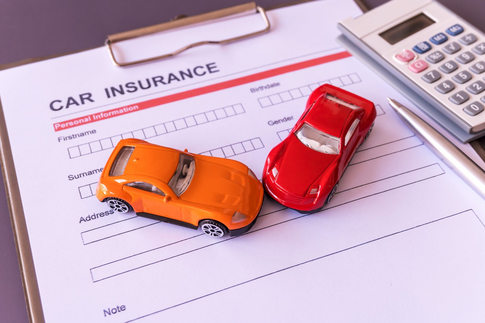 An orange and a red toy car in a collision layout over a printed Car Insurance application form on a clipboard next to a calculator