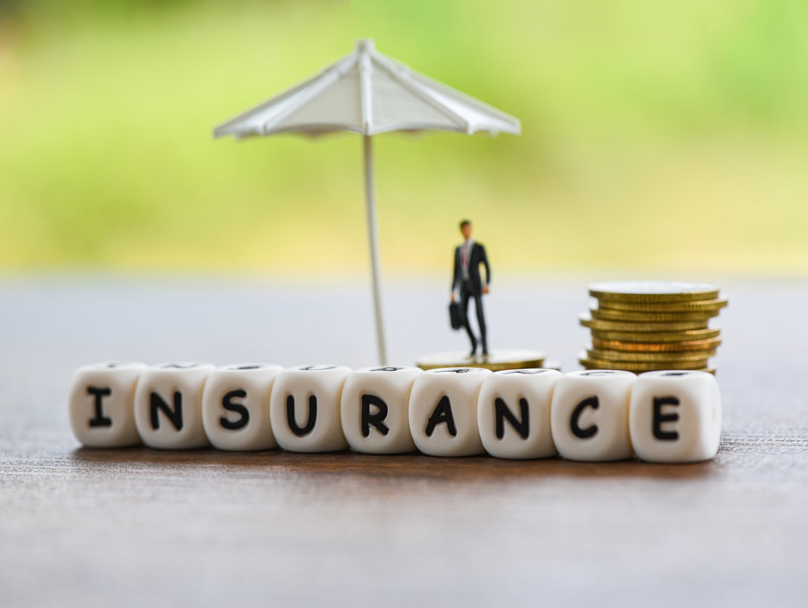 Miniature block letters spelling INSURANCE in front of a small figurine under an umbrella next to stacked coins